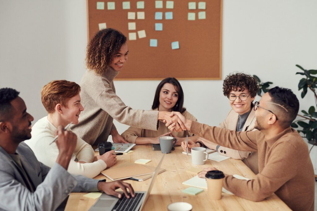 Teammeeting mit mehreren Kollegen an einem Tisch – zwei Personen geben sich die Hand, symbolisieren Zusammenarbeit und erfolgreiche Geschäftspartnerschaft