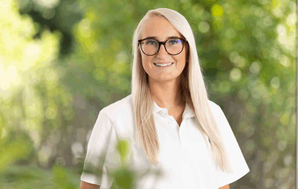 Lächelnde Pflegekraft mit langen blonden Haaren und Brille im weißen Polo steht mit verschränkten Armen vor grünem Naturhintergrund. Symbol für Kompetenz, Vertrauen und persönliche Betreuung in der ambulanten Pflege.